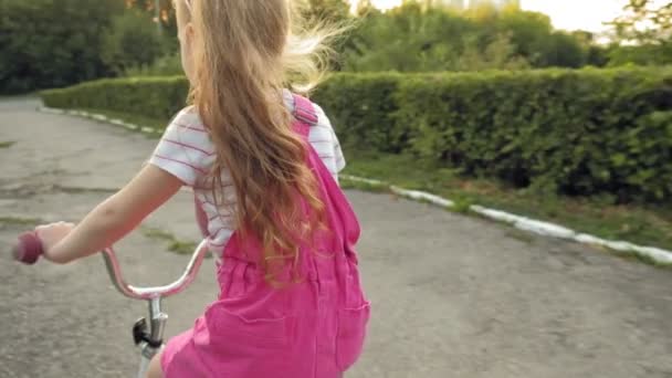 Uma menina feliz, bonita, com longos cabelos loiros em uma saia rosa e jumper monta uma bicicleta infantil na estrada, ela sorri. Movimento super lento — Vídeo de Stock