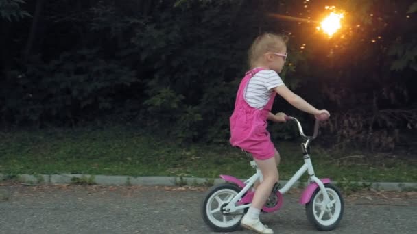 Ein fröhliches, schönes, kleines Mädchen mit langen blonden Haaren in rosa Rock und Pullover fährt mit einem Kinderfahrrad auf der Straße, lächelt sie. Superzeitlupe — Stockvideo