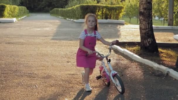 Una niña feliz, hermosa, con el pelo largo y rubio en una falda rosa y saltador monta una bicicleta para niños en la carretera, sonríe. Super cámara lenta — Vídeos de Stock