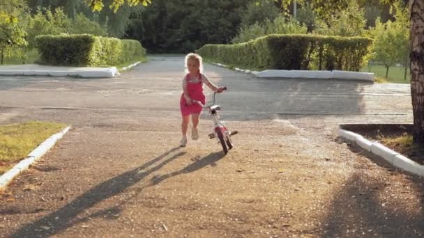 Una niña feliz, hermosa, con el pelo largo y rubio en una falda rosa y saltador monta una bicicleta para niños en la carretera, sonríe. Super cámara lenta — Vídeos de Stock