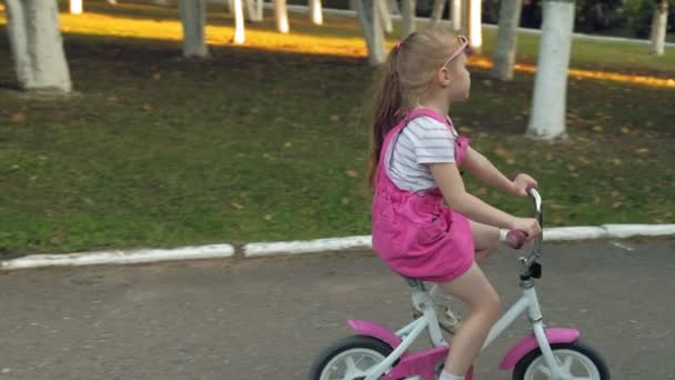 En glad, vacker, liten flicka med långt blont hår i en rosa kjol och jumper rider en barn bike på vägen, hon ler. Super slow motion — Stockvideo