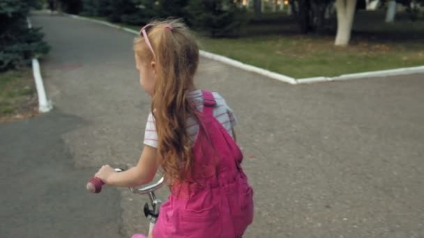 Uzun sarı saç bir pembe etek ve jumper mutlu, güzel, küçük bir kızla yolda o gülümsüyor bir çocuk bisiklet sürmek. Çok yavaş hareket — Stok video
