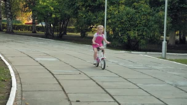 A happy, beautiful, little girl with long blond hair in a pink skirt and jumper rides a childrens bike on the road, she smiles. Super slow motion — Stock Video