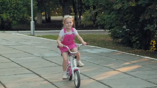 En glad, vacker, liten flicka med långt blont hår i en rosa kjol och jumper rider en barn bike på vägen, hon ler. Super slow motion — Stockvideo