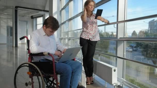 Biznesmen niepełnosprawnych na wózku inwalidzkim w okno z laptopem w pobliżu kobieta korzysta z tabletu — Wideo stockowe