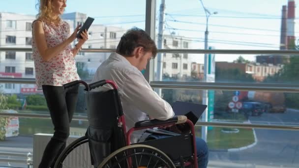 Hombre de negocios discapacitado en una silla de ruedas en una ventana con un ordenador portátil cerca de la mujer utiliza una tableta — Vídeo de stock