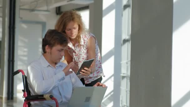 Disabled businessman on a wheelchair at a window with a laptop near woman uses a tablet — Stock Video