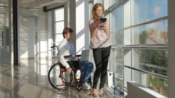 Uomo d'affari disabile su una sedia a rotelle ad una finestra con un computer portatile vicino alla donna utilizza un tablet — Video Stock