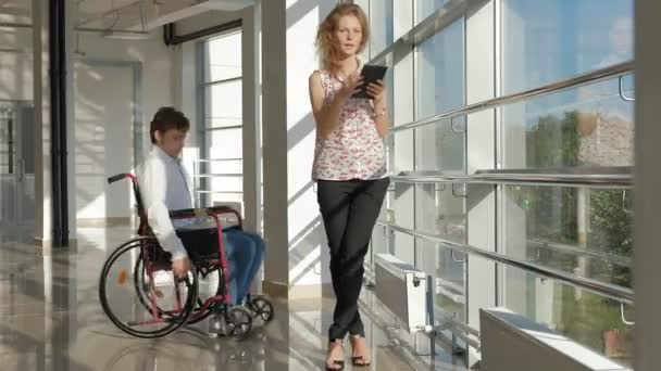 Disabled businessman on a wheelchair at a window with a laptop near woman uses a tablet — Stock Video