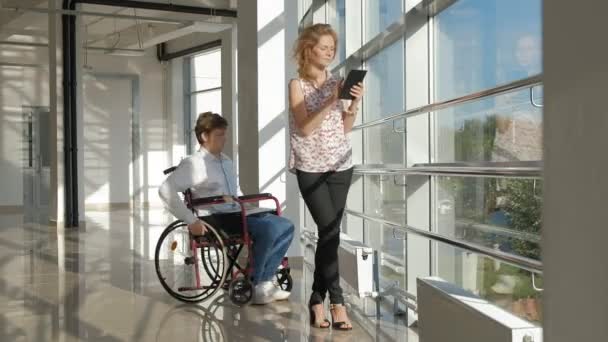 Uomo d'affari disabile su una sedia a rotelle ad una finestra con un computer portatile vicino alla donna utilizza un tablet — Video Stock