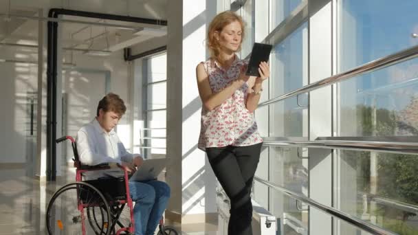 Uomo d'affari disabile su una sedia a rotelle ad una finestra con un computer portatile vicino alla donna utilizza un tablet — Video Stock