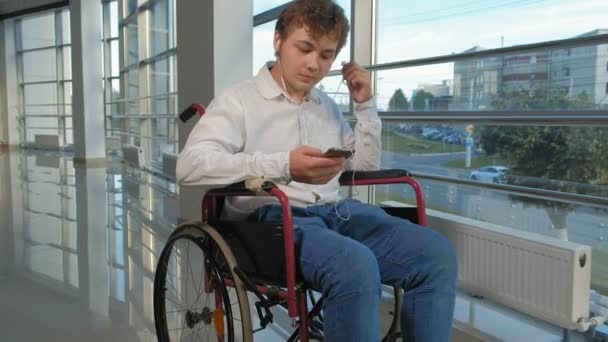 Disabled man on a wheelchair at a window listening to music on headphones from a smartphone — Stock Video