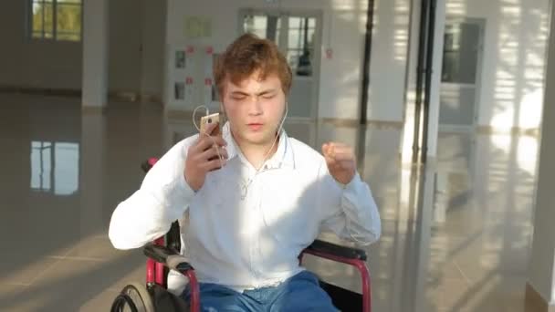 Uomo disabile su una sedia a rotelle ad una finestra che ascolta musica sulle cuffie da uno smartphone — Video Stock