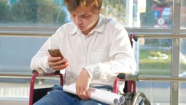 Un homme d'affaires handicapé en fauteuil roulant dans une fenêtre avec un dessin sur une grande feuille de papier, discute du travail par téléphone — Video