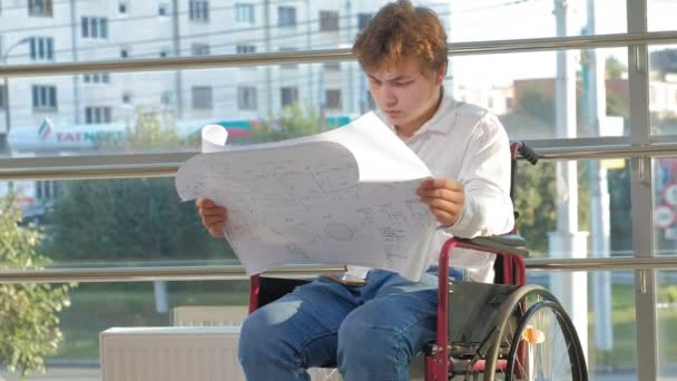 Un uomo d'affari disabile su una sedia a rotelle in una finestra con un disegno su un grande foglio di carta, discute il lavoro per telefono — Video Stock