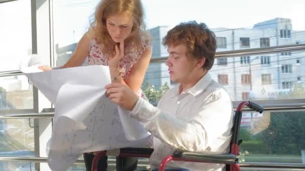 Empresário deficiente em uma cadeira de rodas em uma janela com uma foto em uma grande folha de papel discutindo o trabalho com uma mulher de negócios, discussão no escritório ... — Vídeo de Stock