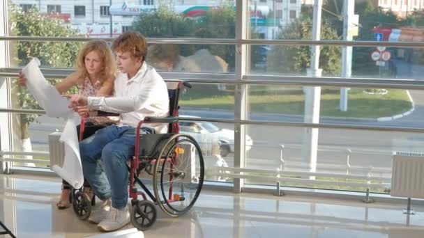 Hombre de negocios discapacitado en una silla de ruedas en una ventana con una imagen en una hoja grande de papel discutir el trabajo con una mujer de negocios, discusión en la oficina ... — Vídeo de stock