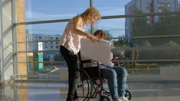 Hombre de negocios discapacitado en una silla de ruedas en una ventana con una imagen en una hoja grande de papel discutir el trabajo con una mujer de negocios, discusión en la oficina ... — Vídeos de Stock