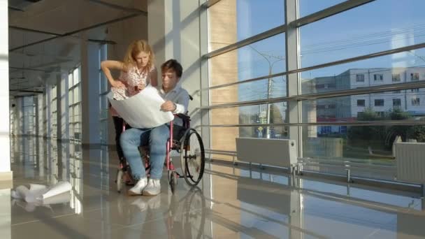 Hombre de negocios discapacitado en una silla de ruedas en una ventana con una imagen en una hoja grande de papel discutir el trabajo con una mujer de negocios, discusión en la oficina ... — Vídeos de Stock