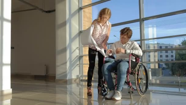 Disabled businessman on a wheelchair in a window with a picture on a large sheet of paper discussing work with a business woman, discussion in the office... — Stock Video