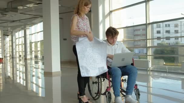 Disabled businessman on a wheelchair in a window with a picture on a large sheet of paper discussing work with a business woman, discussion in the office... — Stock Video