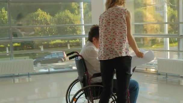 Empresário deficiente em uma cadeira de rodas em uma janela com uma foto em uma grande folha de papel discutindo o trabalho com uma mulher de negócios, discussão no escritório ... — Vídeo de Stock