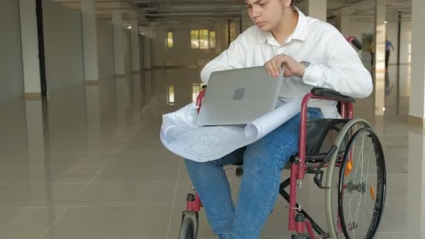 Un uomo d'affari disabile su una sedia a rotelle in una finestra con un disegno su un grande foglio di carta, discute il lavoro per telefono — Video Stock