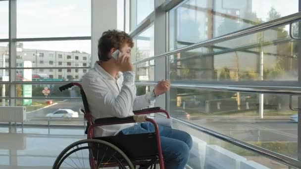 Hombre de negocios discapacitado en una silla de ruedas en una ventana utiliza teléfono inteligente — Vídeos de Stock