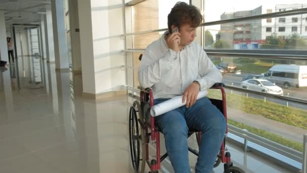 Un hombre de negocios discapacitado en una silla de ruedas en una ventana con un dibujo en una hoja grande de papel, discute el trabajo por teléfono — Vídeo de stock