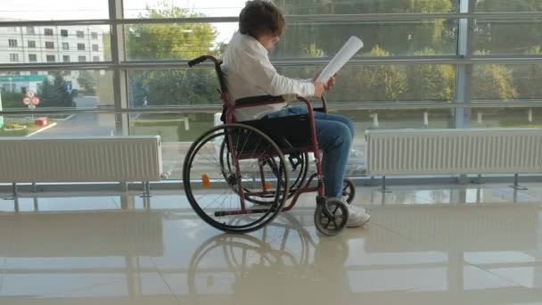 Een handicap zakenman op een rolstoel in een venster met een tekening op een groot vel papier, bespreekt werk per telefoon — Stockvideo