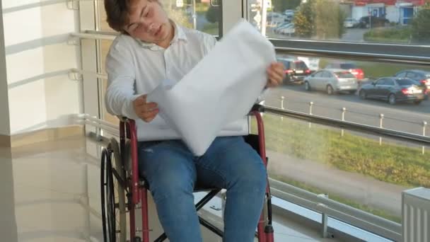 Un uomo d'affari disabile su una sedia a rotelle in una finestra con un disegno su un grande foglio di carta, discute il lavoro per telefono — Video Stock