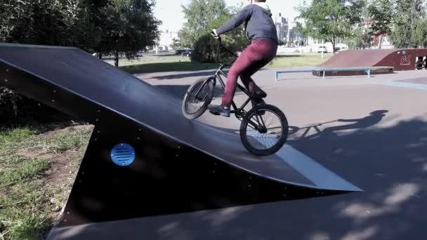 Un niño está montando trucos de ciclismo BMX en un parque de skate en un día soleado. Super cámara lenta — Vídeos de Stock