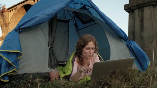 Junge Frau arbeitet hinter einem Laptop in einem Touristenzelt nahe der alten Brücke — Stockvideo