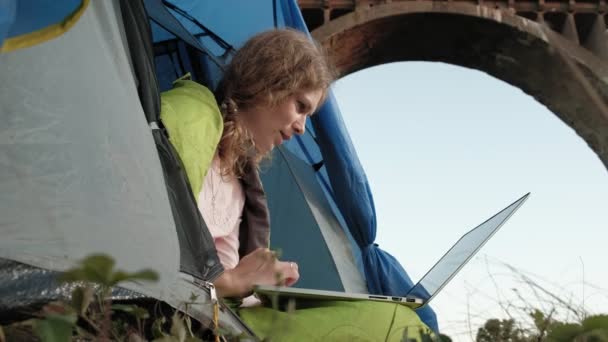 Mujer joven que trabaja detrás de un ordenador portátil en una tienda de campaña turística cerca del viejo puente — Vídeos de Stock