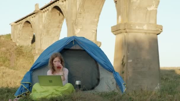 Jovem mulher trabalhando atrás de um laptop em uma tenda turística perto da ponte velha — Vídeo de Stock