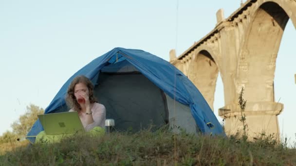 Jovem mulher trabalhando atrás de um laptop em uma tenda turística perto da ponte velha — Vídeo de Stock