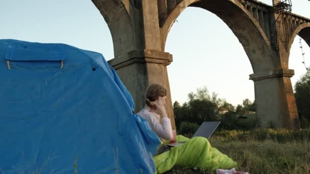 Jovem mulher trabalhando atrás de um laptop em uma tenda turística perto da ponte velha — Vídeo de Stock