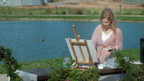 A pretty woman with red hair, paints a picture on canvas, which stands on the easel. The lady is in the open air near the lake of the river, she draws from life — Stock Video