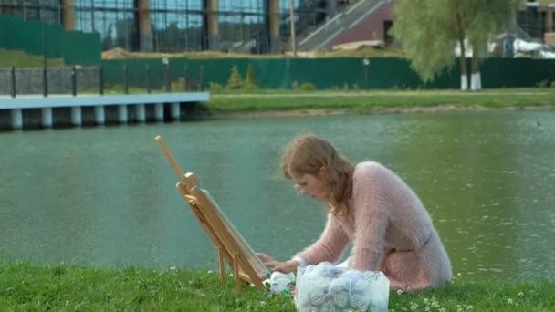 En vacker kvinna med rött hår, målar upp en bild på duk, som står på staffliet. Damen är i öppna luften nära sjön av floden, hon drar från liv — Stockvideo