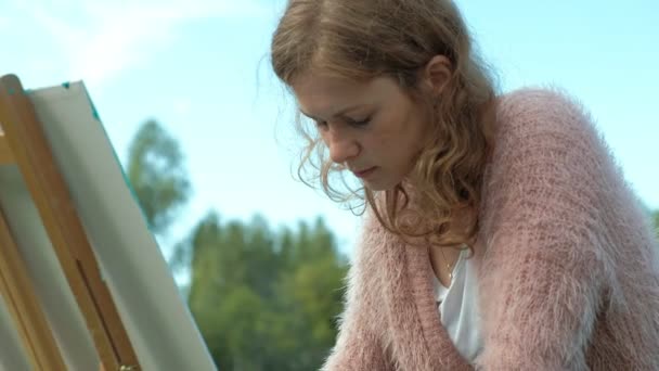 Een mooie vrouw met rood haar, schetst een beeld op doek, dat op de ezel staat. De dame is in de open lucht in de buurt van het meer van de rivier, ze tekent uit het leven — Stockvideo