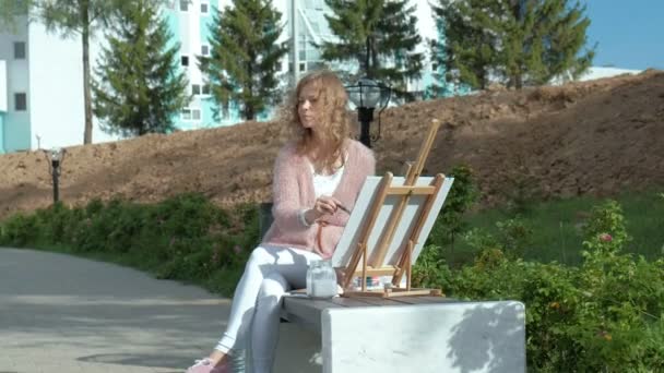 Uma mulher bonita com cabelo ruivo, pinta um quadro em tela, que fica no cavalete. A senhora está ao ar livre perto do lago do rio, ela desenha da vida — Vídeo de Stock