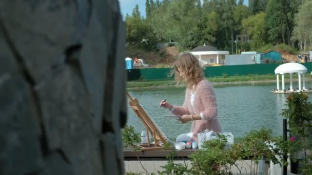 Uma mulher bonita com cabelo ruivo, pinta um quadro em tela, que fica no cavalete. A senhora está ao ar livre perto do lago do rio, ela desenha da vida — Vídeo de Stock