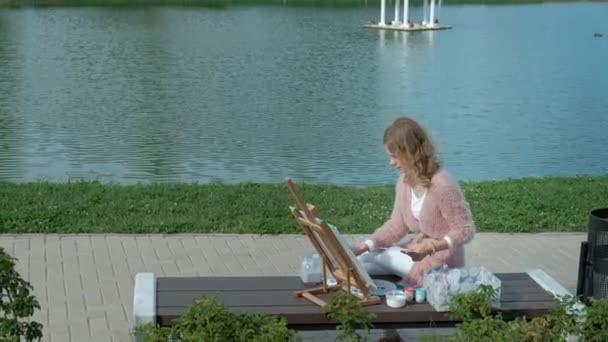 Een mooie vrouw met rood haar, schetst een beeld op doek, dat op de ezel staat. De dame is in de open lucht in de buurt van het meer van de rivier, ze tekent uit het leven — Stockvideo