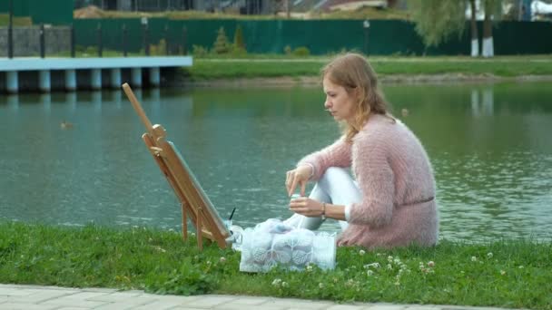 A pretty woman with red hair, paints a picture on canvas, which stands on the easel. The lady is in the open air near the lake of the river, she draws from life — Stock Video