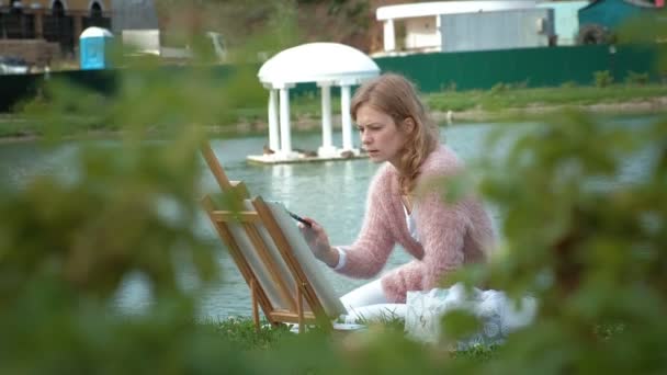 Een mooie vrouw met rood haar, schetst een beeld op doek, dat op de ezel staat. De dame is in de open lucht in de buurt van het meer van de rivier, ze tekent uit het leven — Stockvideo