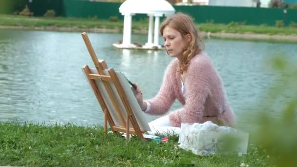 A pretty woman with red hair, paints a picture on canvas, which stands on the easel. The lady is in the open air near the lake of the river, she draws from life — Stock Video