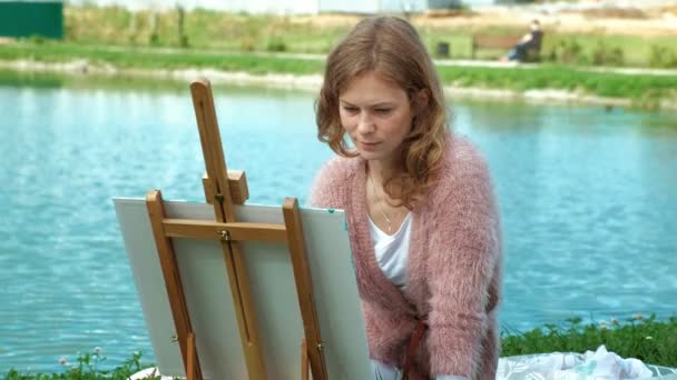 Uma mulher bonita com cabelo ruivo, pinta um quadro em tela, que fica no cavalete. A senhora está ao ar livre perto do lago do rio, ela desenha da vida — Vídeo de Stock