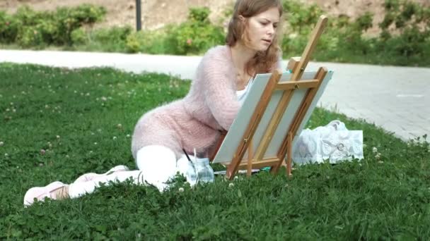Una mujer bonita con el pelo rojo, pinta un cuadro sobre lienzo, que se encuentra en el caballete. La señora está en el aire libre cerca del lago del río, ella saca de la vida — Vídeo de stock