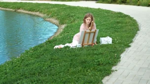 A pretty woman with red hair, paints a picture on canvas, which stands on the easel. The lady is in the open air near the lake of the river, she draws from life — Stock Video