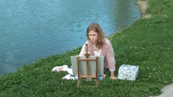 En vacker kvinna med rött hår, målar upp en bild på duk, som står på staffliet. Damen är i öppna luften nära sjön av floden, hon drar från liv — Stockvideo
